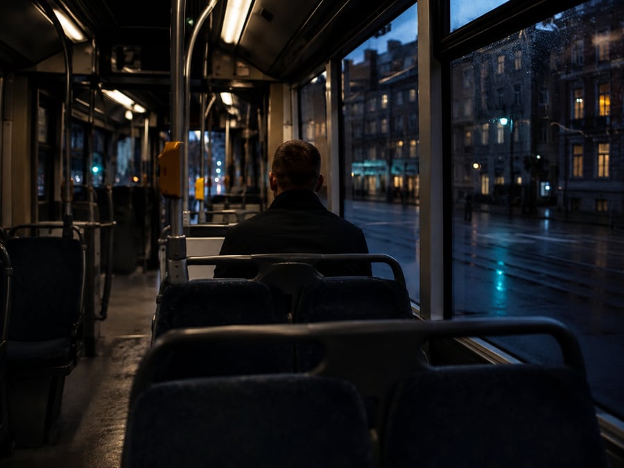 A lone passenger seen from behind on a tram at night, city lights through the wet window.